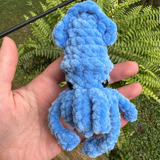 A small blue fuzzy lobster-shaped plush toy or accessory being held in a person's hand outdoors, with green ferns and a wooden surface in the background.