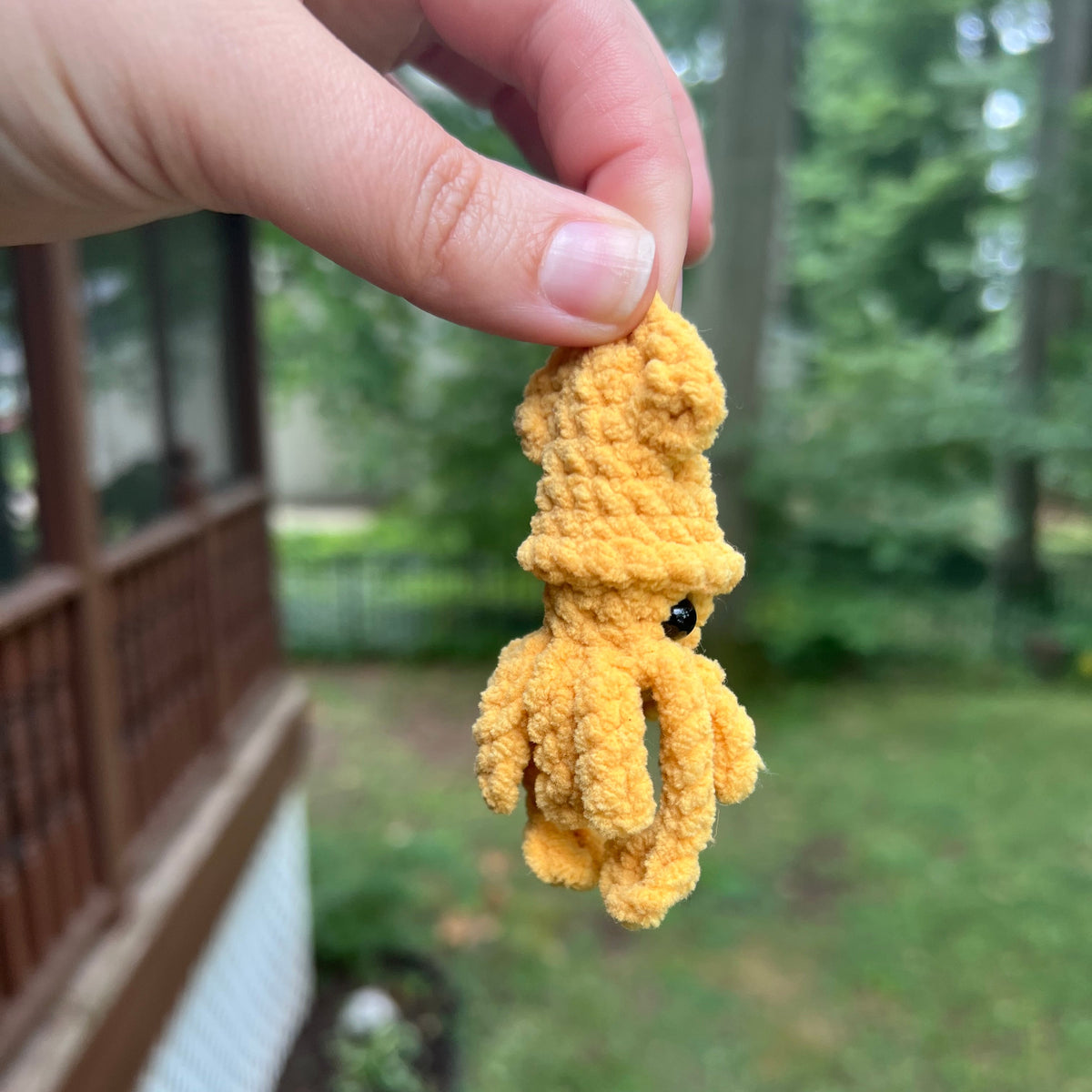 A hand holding a small yellow textured toy or knot against a blurred outdoor background with greenery and a wooden structure.