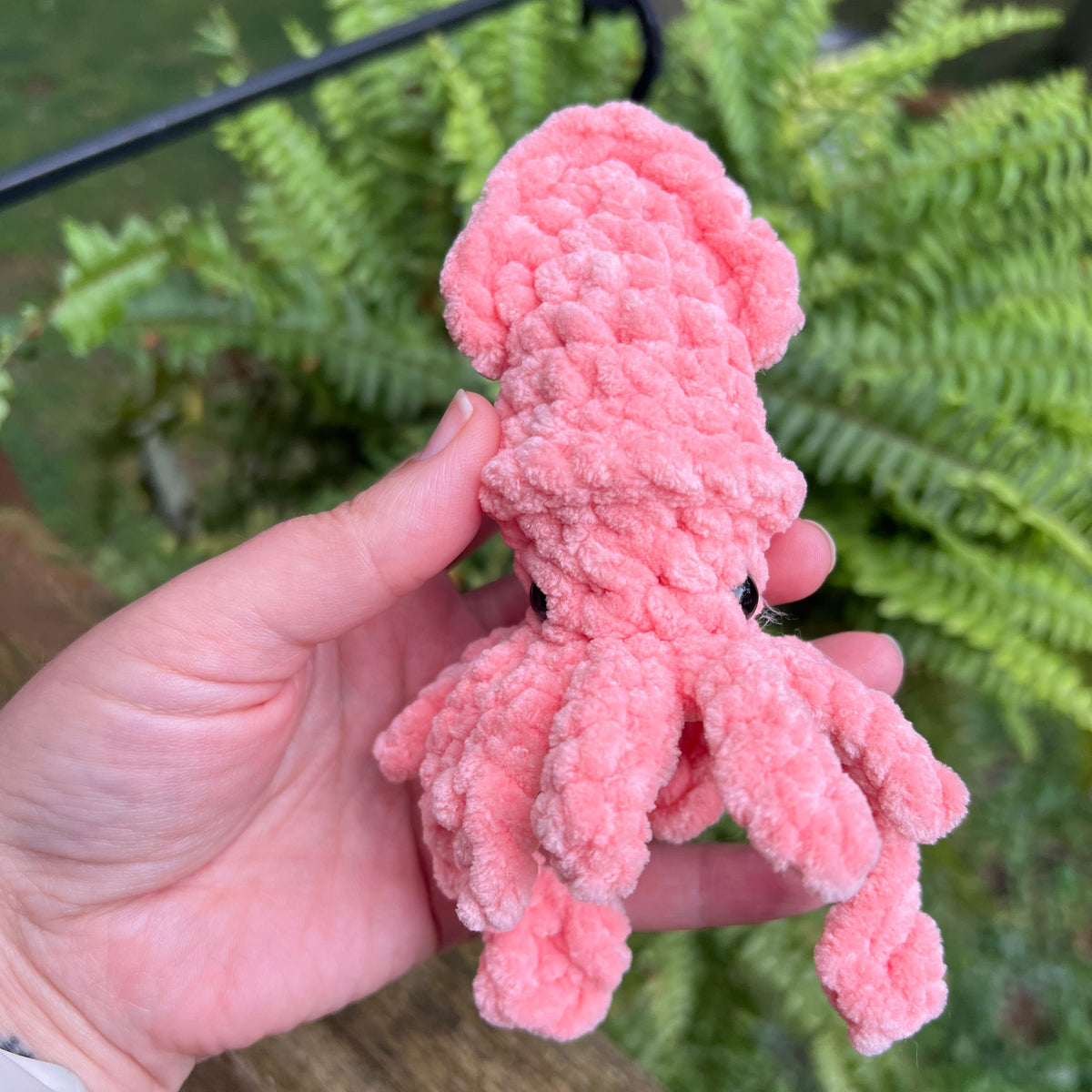 A pink fuzzy octopus plush toy being held in a person's hand, with green ferns and a wooden fence in the background.