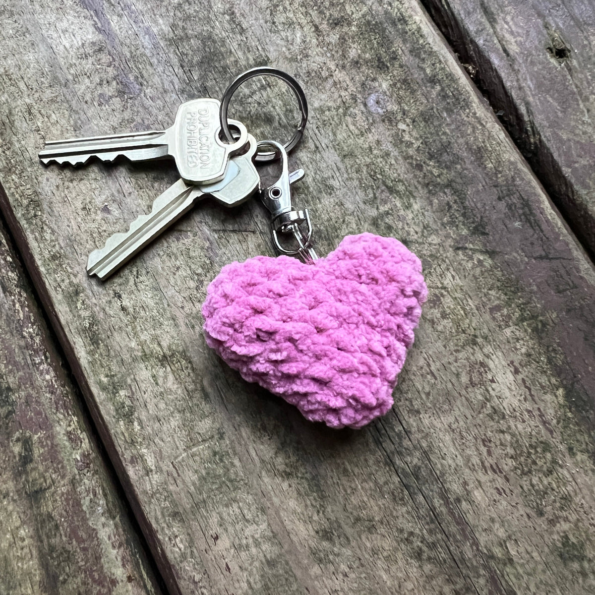 A pink crocheted heart-shaped keychain with silver metal keys and clasp, resting on a weathered gray wooden plank surface.