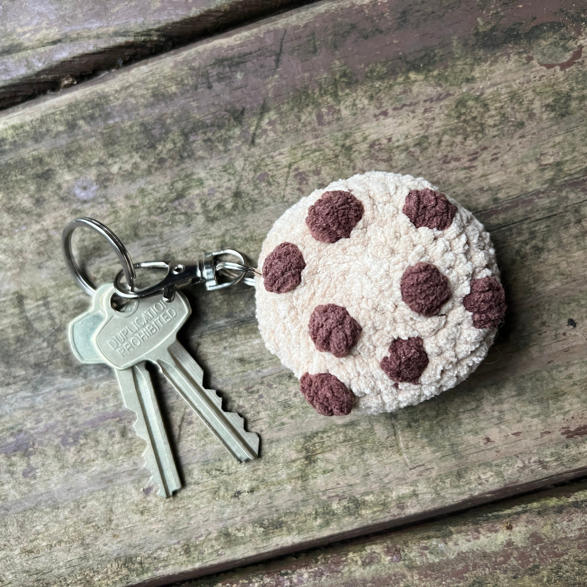 A small chocolate chip cookie, attached to a metal keyring with two keys, placed on a weathered wooden surface.