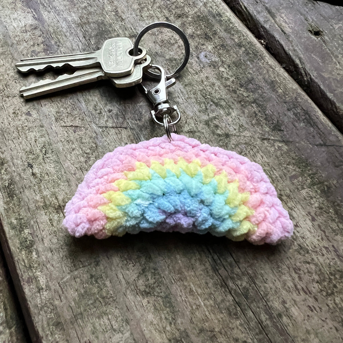A crocheted rainbow-shaped keychain with a metal key ring and clasp, resting on a weathered wooden surface. The keychain features soft pastel stripes in pink, yellow, light blue, and lavender.