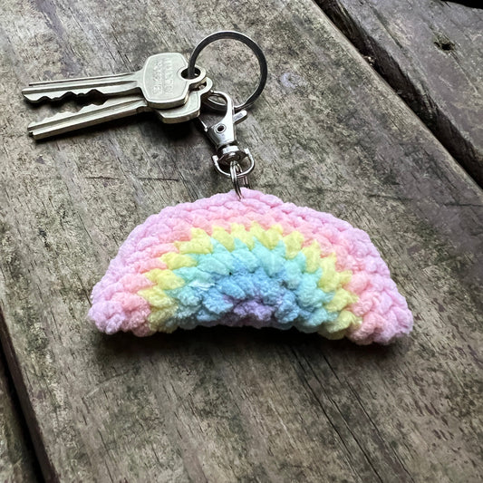 A crocheted rainbow-shaped keychain with a metal key ring and clasp, resting on a weathered wooden surface. The keychain features soft pastel stripes in pink, yellow, light blue, and lavender.