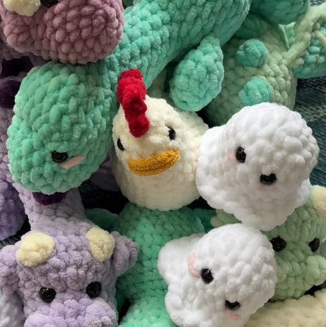 A pile of handmade crocheted plush toys, including a mint green dinosaur, a purple dinosaur, a white duck, and several white fluffy ghost characters, arranged together on a patterned fabric background.